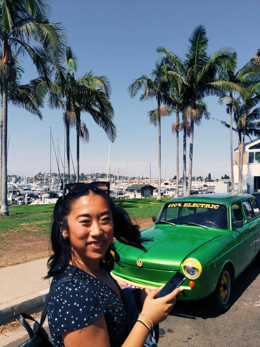 A smiling woman stands in front of a bright green vintage car with “100% ELECTRIC” on the windshield, surrounded by palm trees and a marina with boats in the background on a sunny day.