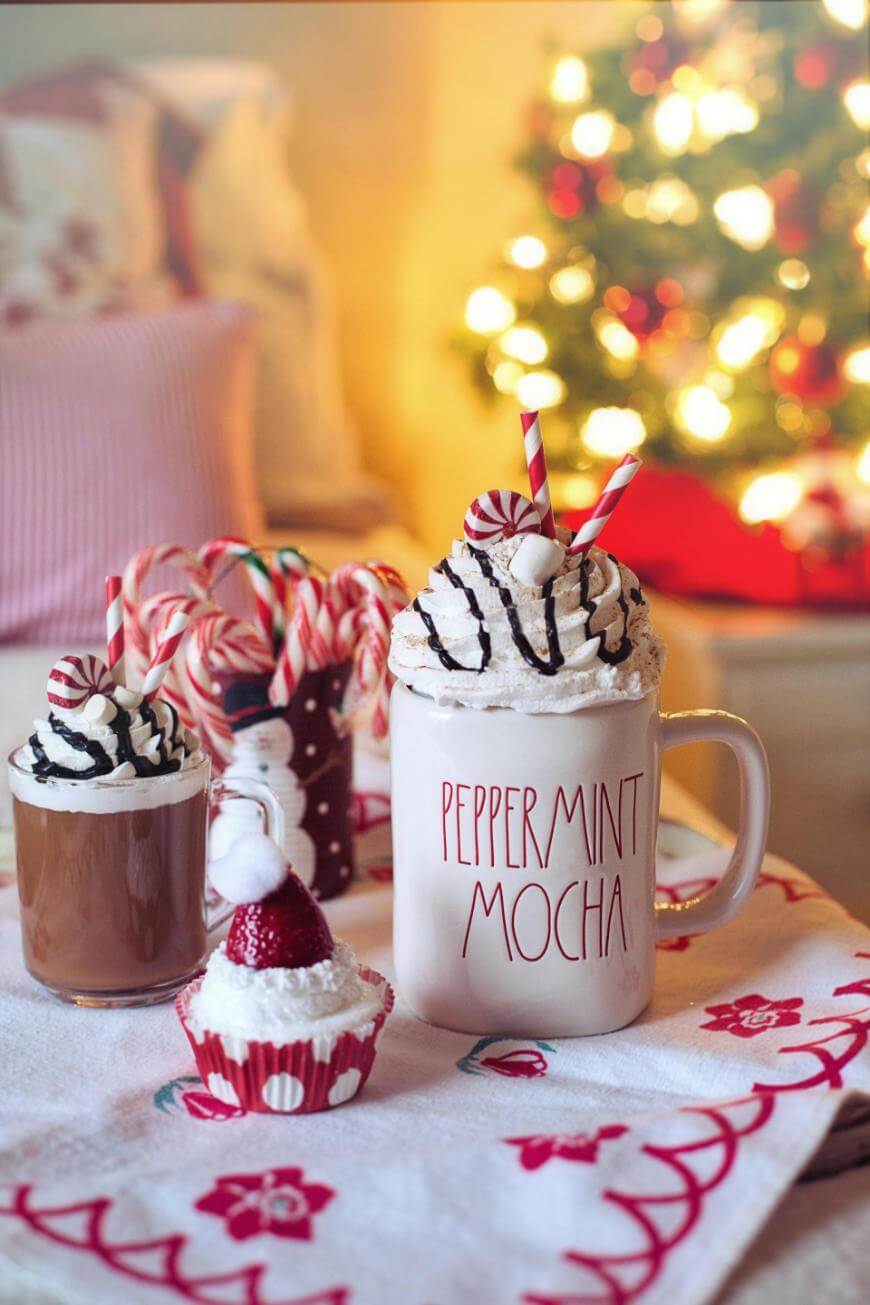 A festive scene with a mug labeled “Peppermint Mocha” filled with whipped cream, chocolate drizzle, and candy canes, next to a small cup, a cupcake, and a decorated tree glowing in the background.