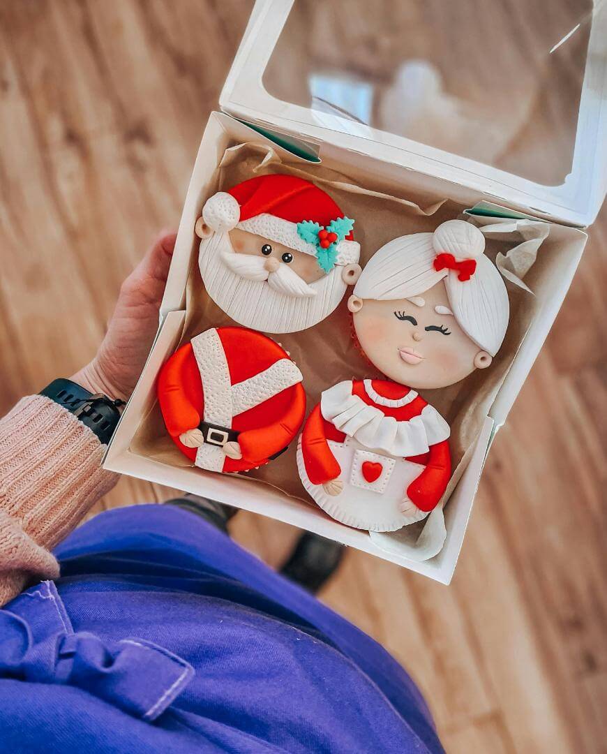 A hand holds an open box with three decorated cookies: one shaped like Santa Claus, one like Mrs. Claus, and one showing Santa’s suit, all with festive icing details.