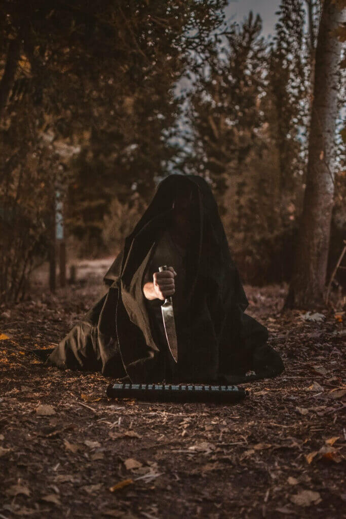 A person in a dark cloak kneels on forest ground, holding a large knife upright. The figure is mostly in shadow, with trees and fallen leaves surrounding them, and an object with buttons or keys lies in front.