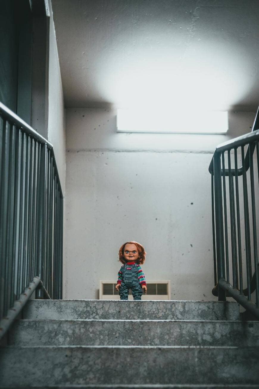 A small Chucky doll stands at the top of a concrete staircase in a dimly lit, industrial setting, with a bright fluorescent light on the wall above.