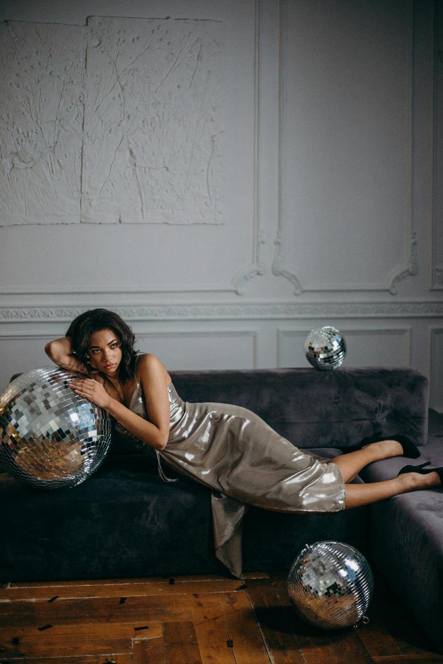 A woman in a shiny, silver dress lounges on a dark velvet sofa with one arm around a large disco ball. Two more disco balls sit on the floor. The room has ornate white walls and wooden flooring.