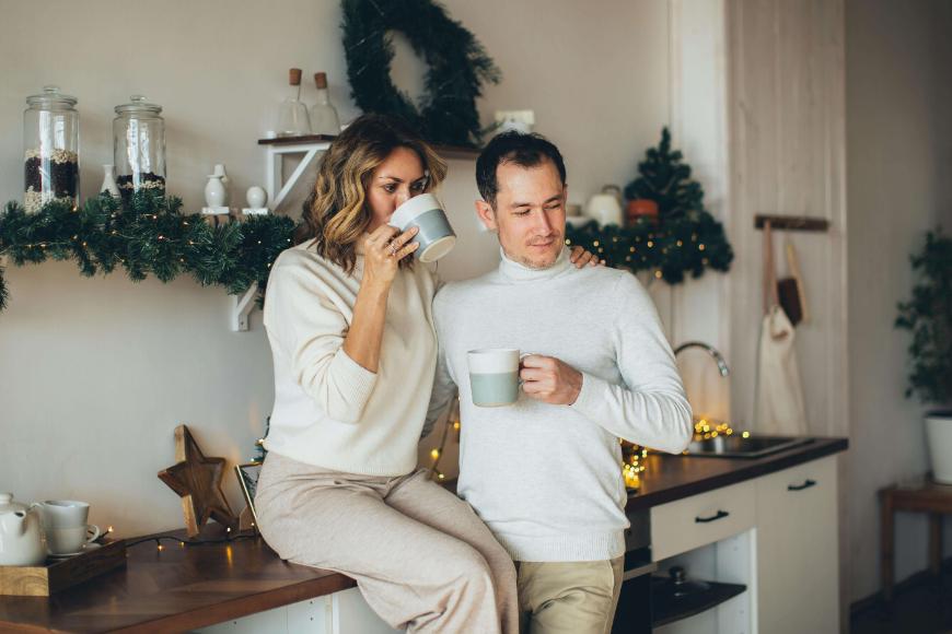 A woman sitting on a kitchen counter and a man standing beside her, both wearing light sweaters and holding mugs. The kitchen is decorated with garlands and festive lights, creating a cozy holiday atmosphere.