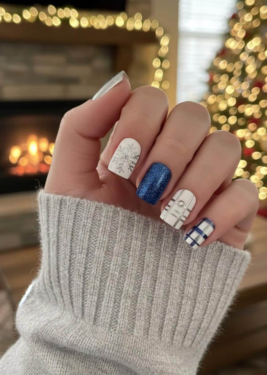 A hand with festive winter-themed nail art, featuring blue glitter, snowflakes, and plaid designs, is held up in front of a fireplace and a lit Christmas tree. The person is wearing a light gray knit sweater.