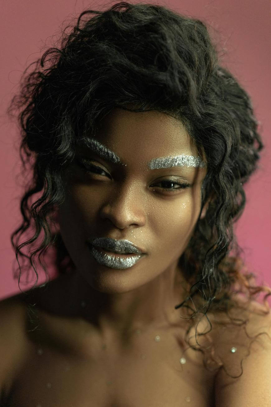 A person with dark, wavy hair poses against a pink background. They have silver glitter on their eyebrows and lips, and small silver sparkles on their skin. The expression is calm and direct.