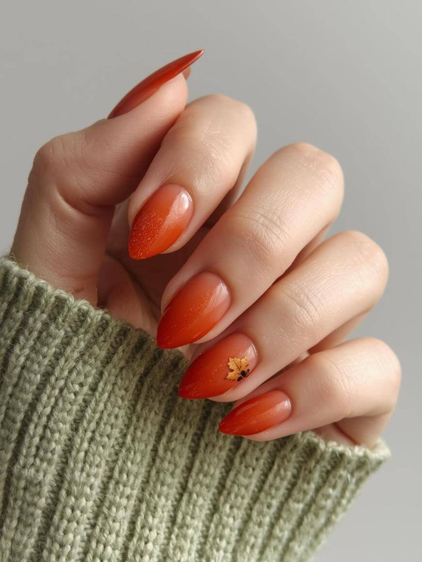 A hand with long, almond-shaped nails painted in a gradient orange, featuring a small gold autumn leaf design on the ring finger. The person is wearing a green knitted sweater.