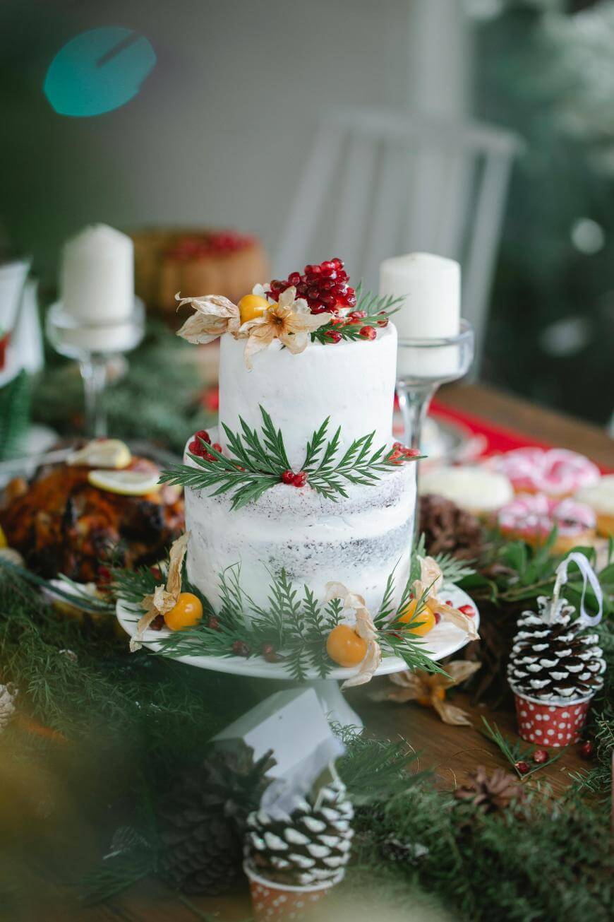 A two-tiered white cake decorated with green leaves, red berries, and yellow fruits sits on a festive table surrounded by candles, pinecones, and holiday greenery.