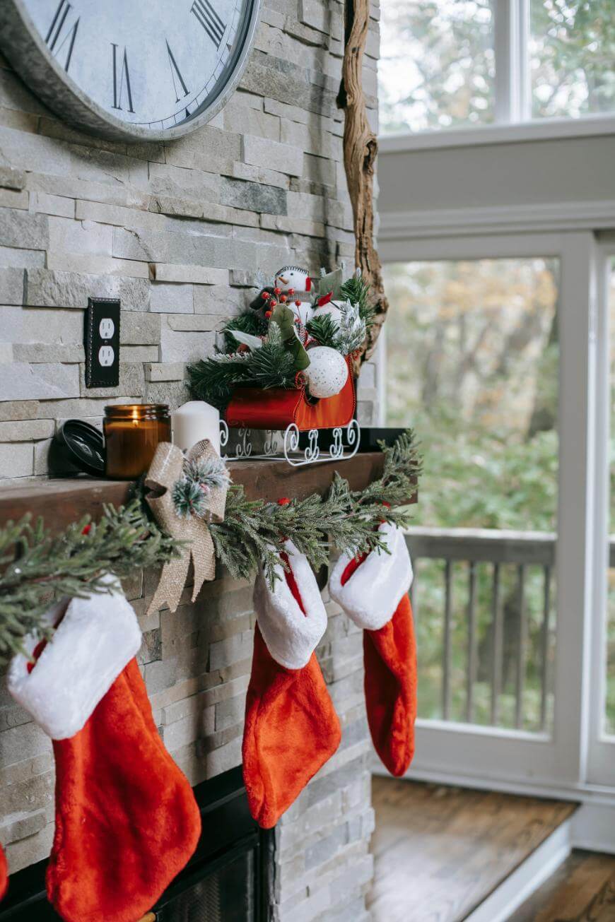 A fireplace mantel decorated with red and white Christmas stockings, garland, and holiday ornaments. A snowman figurine, a candle, and festive greenery are displayed on the mantel. A large clock hangs above.