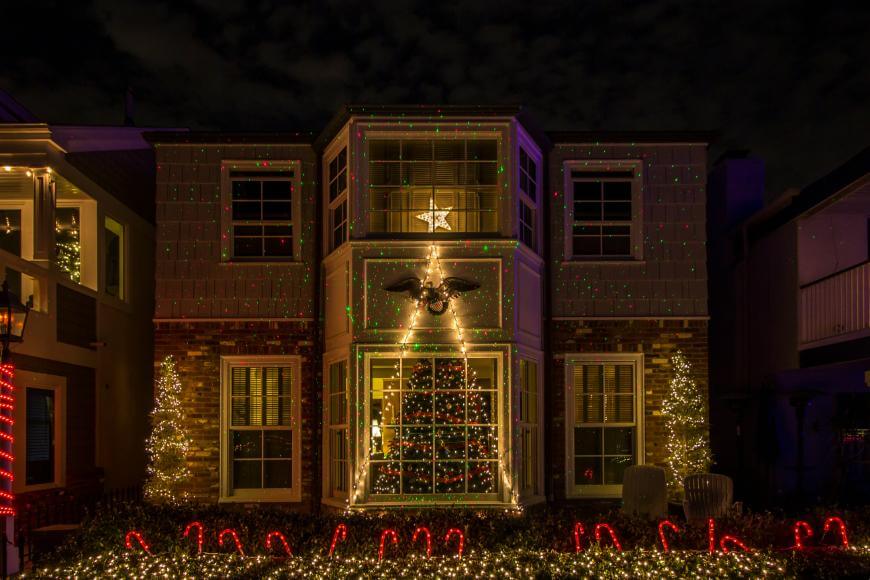 A house decorated with Christmas lights, including a lit star above the door, two illuminated trees, a Christmas tree visible through the window, and red and white candy cane lights lining the walkway.
