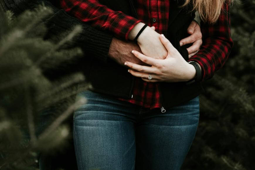 A close-up of two people standing among evergreen trees, with one person embracing the other from behind. Both wear casual clothes; the front person wears a red plaid shirt and jeans, showing a ring on their finger.