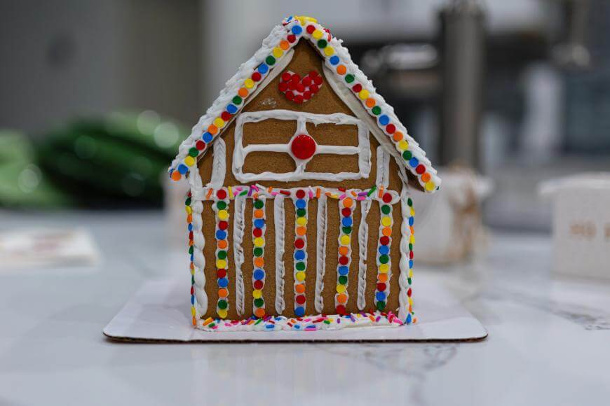 A decorated gingerbread house with colorful candy dots, white icing, a red heart, and a red candy above the door, displayed on a white surface.