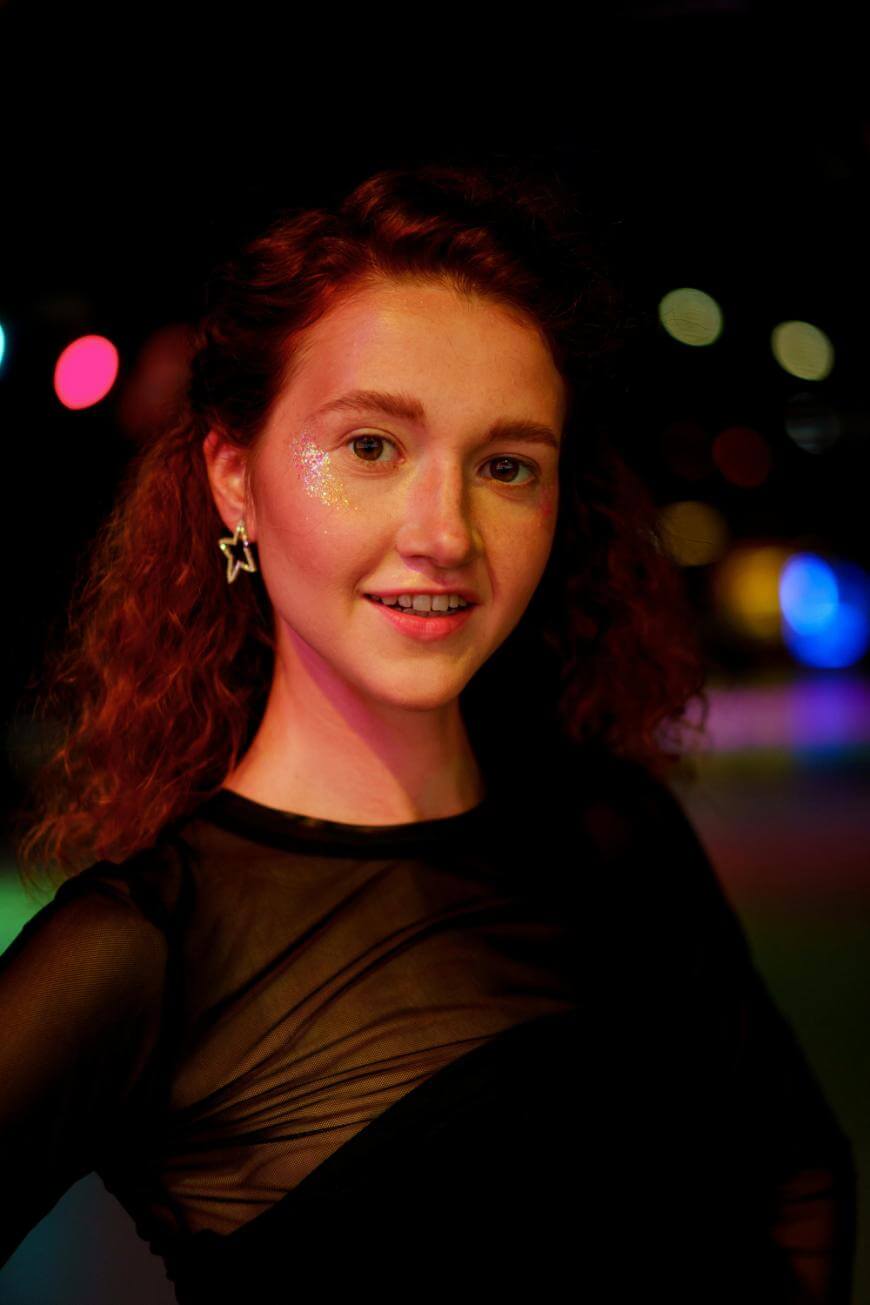 A young woman with curly red hair, wearing a sheer black top and star-shaped earrings, smiles at the camera. She has glitter on her cheek and colorful lights blur in the background.