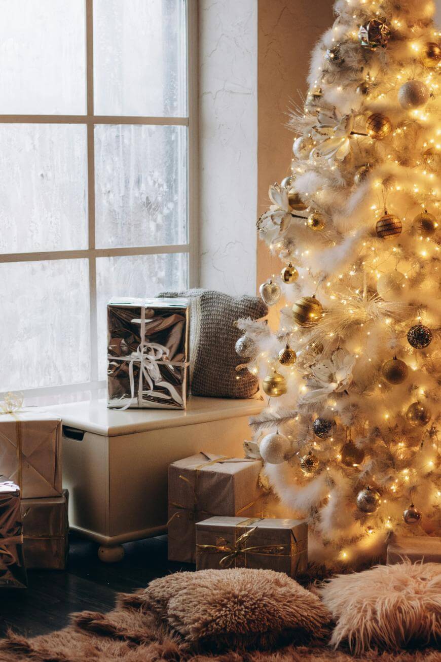 A white Christmas tree decorated with gold ornaments and warm lights stands by a frosted window, surrounded by gold-wrapped presents and cozy pillows on the floor.