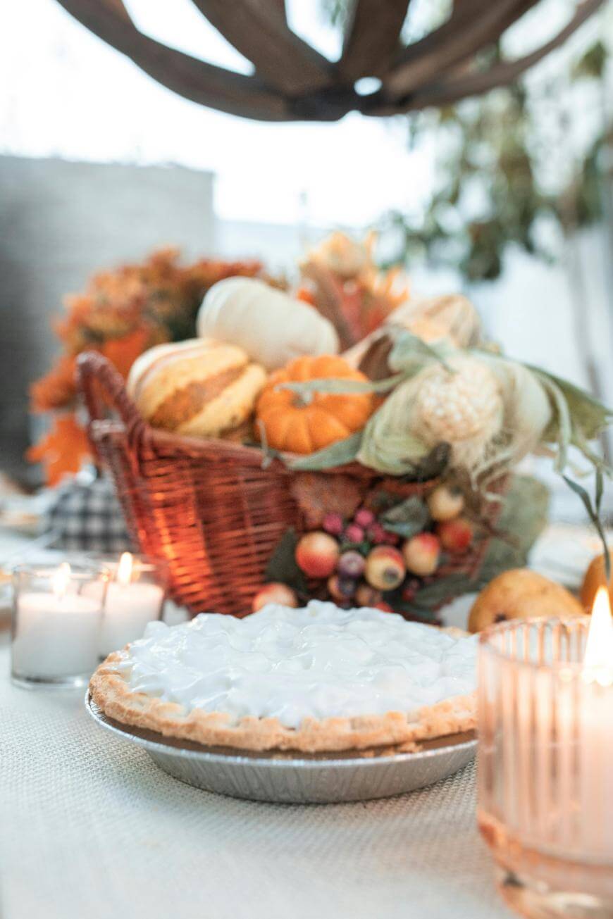 A pie with a meringue topping sits on a table decorated with lit candles and a basket filled with pumpkins, gourds, and autumn leaves, creating a festive fall atmosphere.