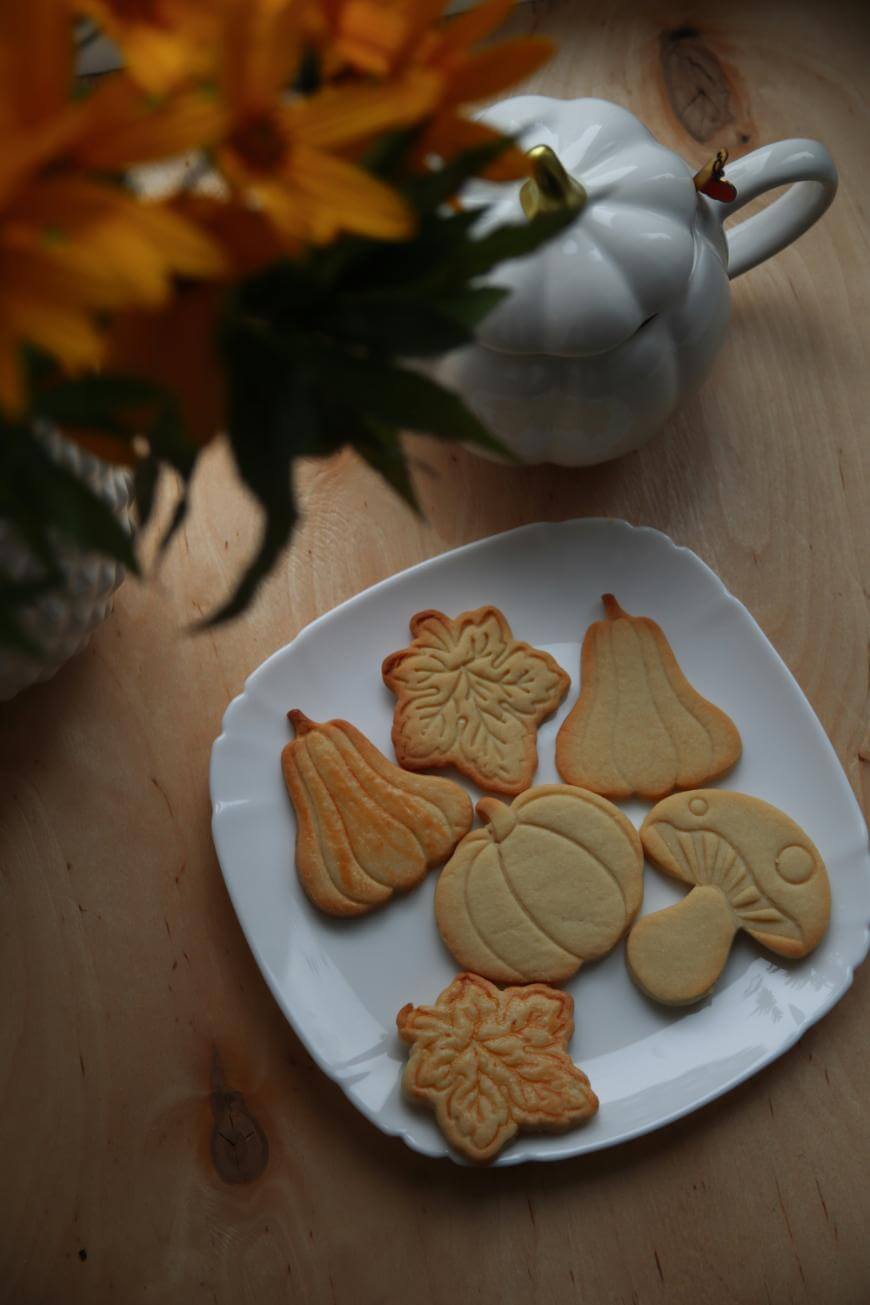 A white plate with autumn-themed cookies shaped like leaves, pumpkins, and gourds sits on a wooden table next to a white pumpkin-shaped teapot. Yellow flowers are partially visible in the top left corner.