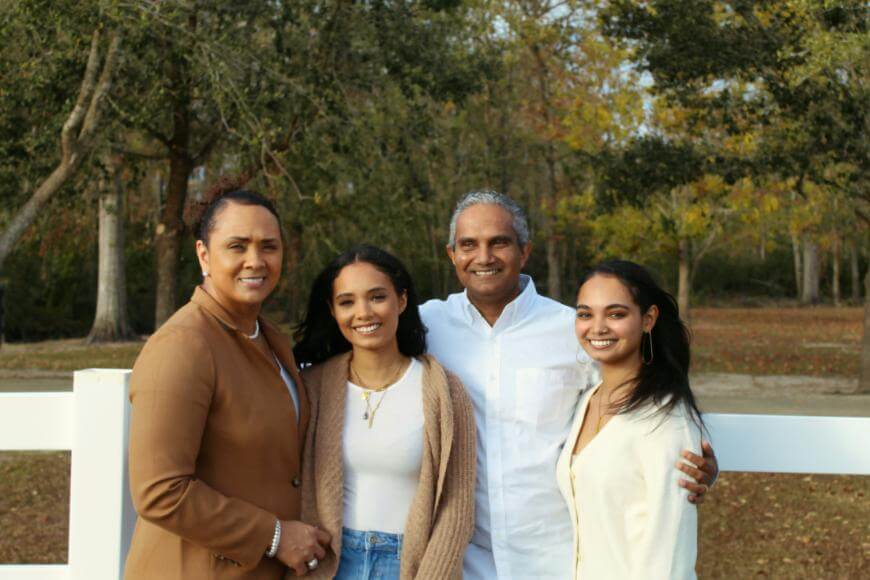 A family of four stands outdoors in front of a white fence and trees. Two women and two young women smile at the camera, dressed in neutral fall clothing. The background shows autumn foliage and a grassy area.