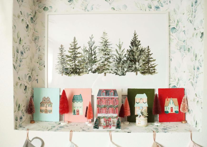 A shelf decorated with miniature house and tree ornaments, colorful holiday cards, and a framed painting of snow-covered pine trees. The background features a leafy wallpaper.