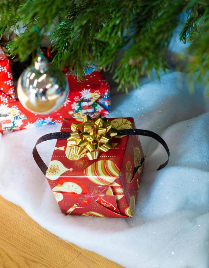 A red gift box with gold holiday designs and a shiny gold bow sits under a Christmas tree, next to a silver ornament and artificial snow. Another wrapped present is partially visible in the background.