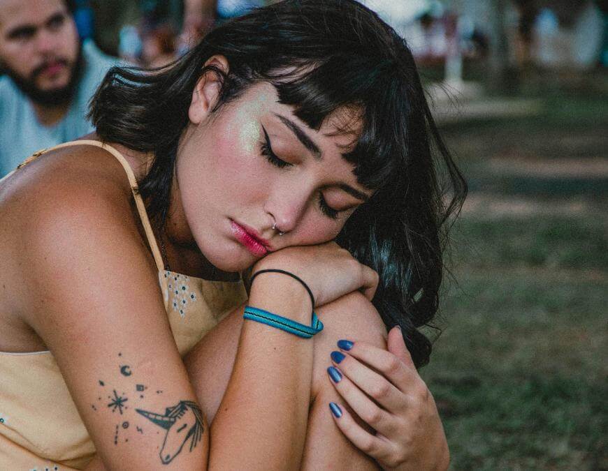 A young woman with dark hair and a nose ring rests her head on her knees outdoors. She has glitter makeup, blue nail polish, a unicorn tattoo, and closed eyes, appearing thoughtful or tired. A blurred person is in the background.