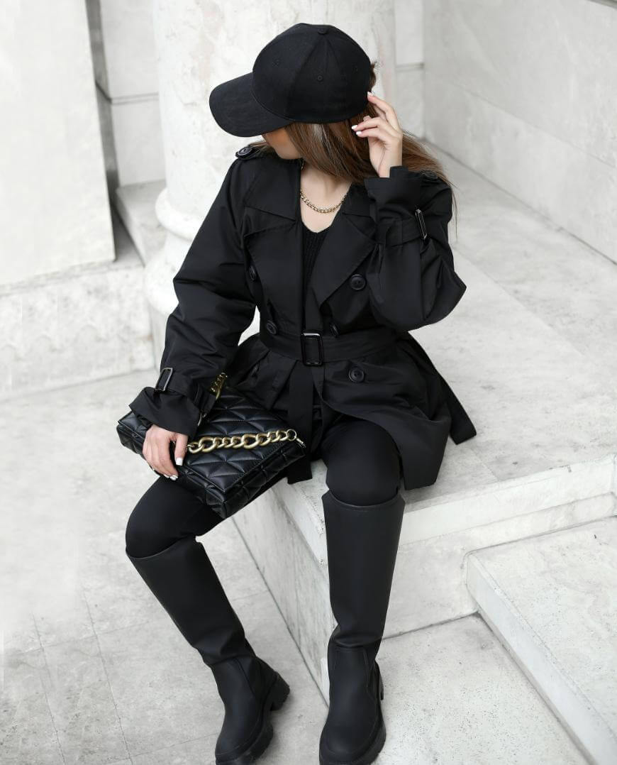 A person wearing a black cap, trench coat, leggings, and boots sits on white stone steps, holding a quilted black handbag with a gold chain, facing away from the camera.
