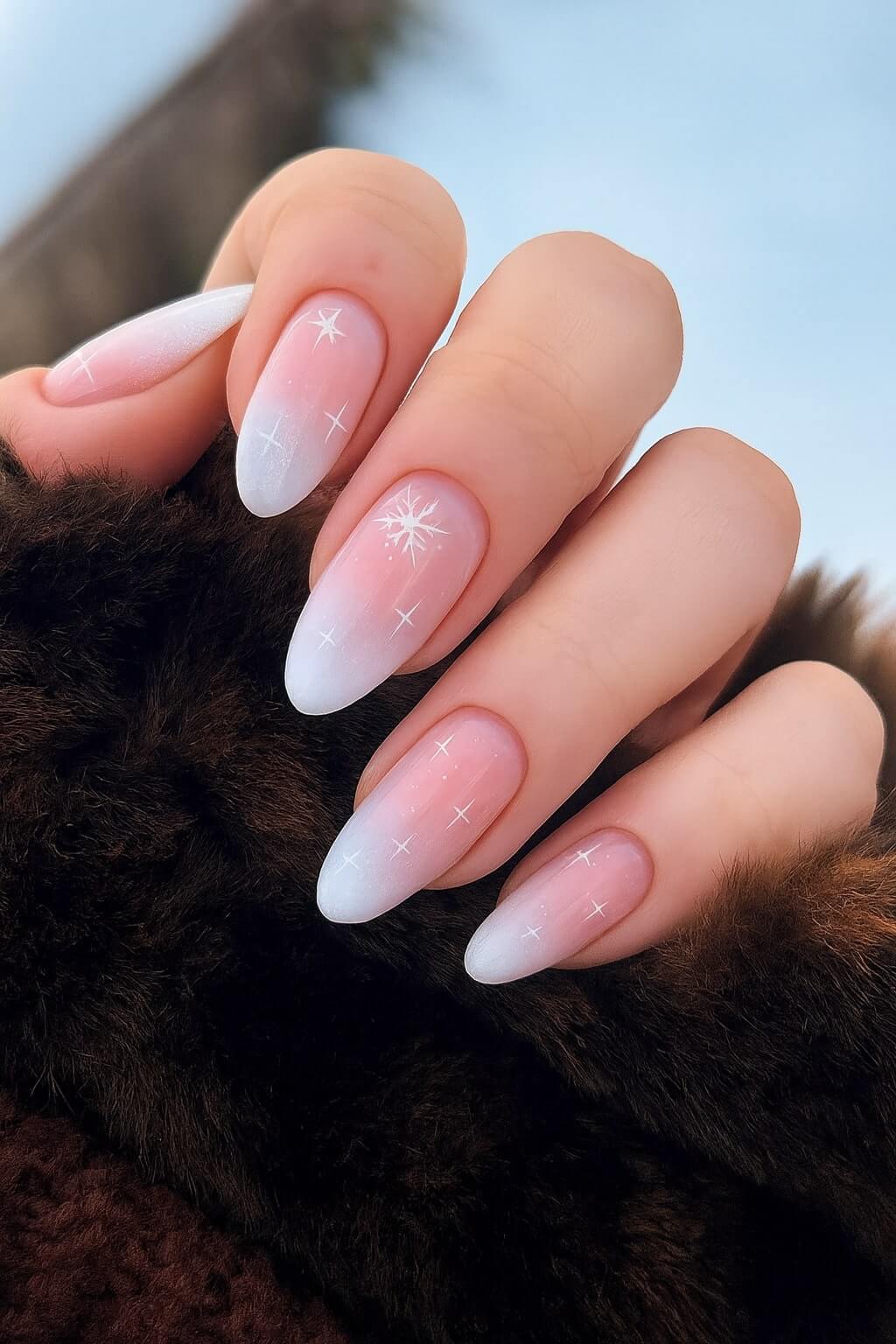 A hand with almond-shaped nails featuring a soft pink to white ombre design, decorated with delicate white star and cross patterns, resting on a dark brown fur fabric.