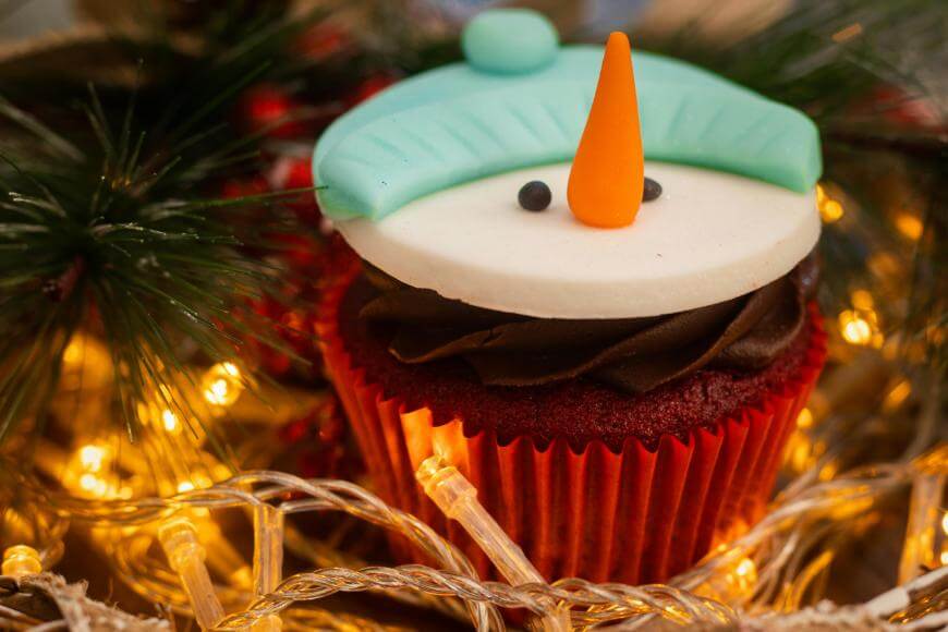 A festive cupcake decorated to look like a snowman’s face with a blue hat, orange carrot nose, and chocolate frosting, surrounded by pine branches and warm holiday string lights.