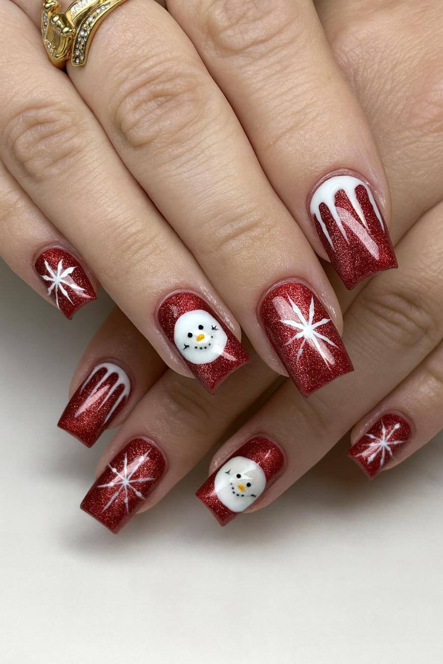 A hand with red glittery Christmas-themed nails featuring snowflake designs, two nails with white snowman faces, and two nails with white icicle patterns near the cuticles.