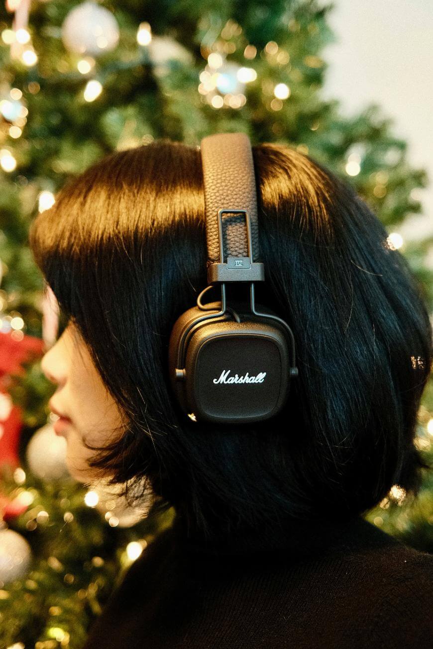 A person with short black hair wears black Marshall headphones in front of a decorated Christmas tree with lights and ornaments.