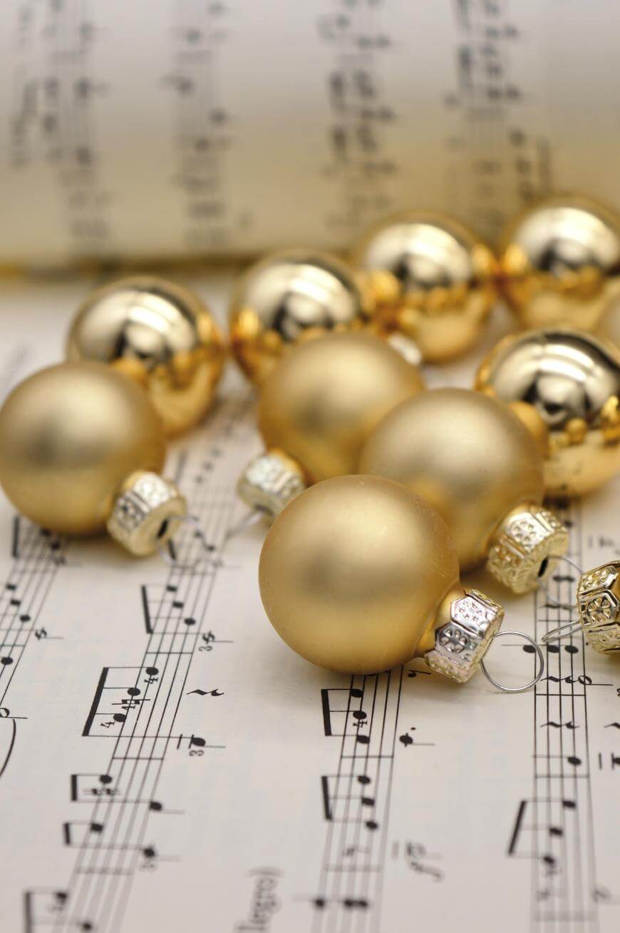 Gold Christmas ornaments rest on sheets of music with musical notes and staff lines visible; more ornaments and a rolled-up sheet of music are blurred in the background.