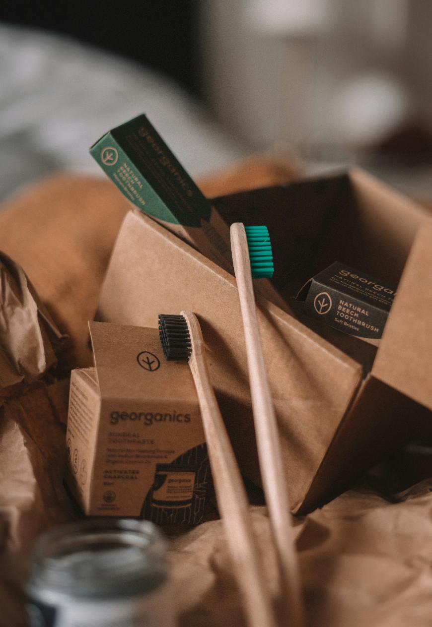 A cardboard box contains eco-friendly Georgians products, including two bamboo toothbrushes—one with green bristles and one with black bristles—and packaging for natural toothpaste. The scene is set on crumpled brown paper.