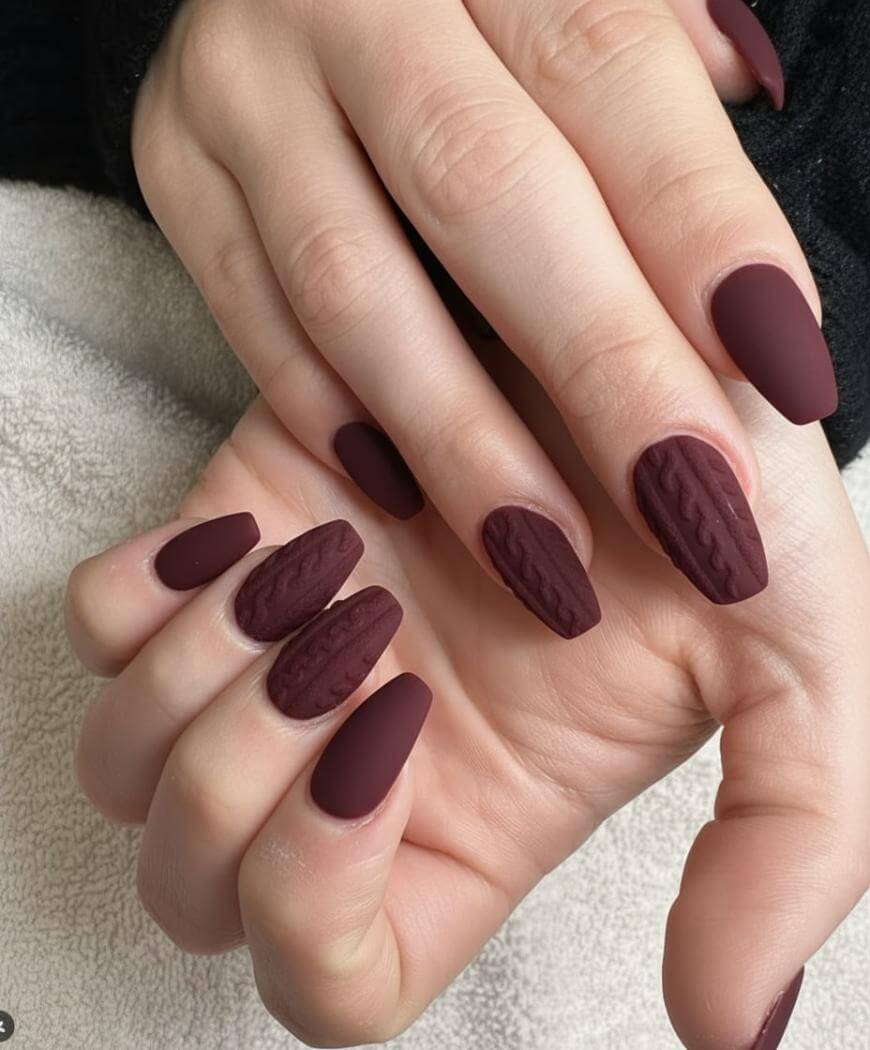 A close-up of hands with matte dark burgundy almond-shaped nails; some nails feature a textured knit-like pattern accent.