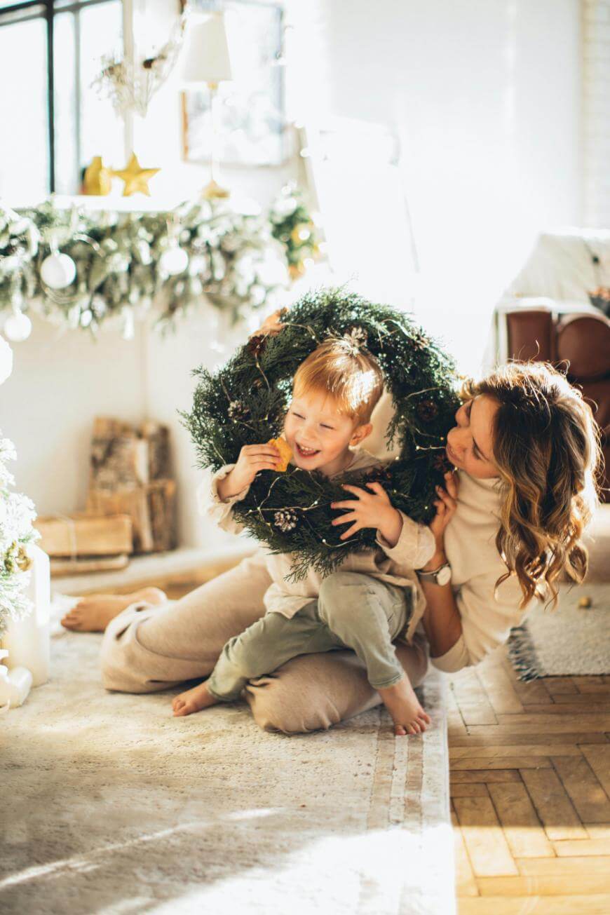 A woman and a young boy sit on the floor in a cozy, decorated room. The boy smiles while holding a large Christmas wreath around his head, and the woman lovingly hugs him from behind.