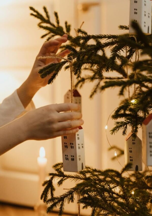 A person hangs a house-shaped ornament on a Christmas tree decorated with lights. A lit candle glows softly in the background, creating a warm, festive atmosphere.