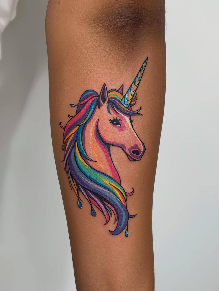 A colorful tattoo of a unicorn head with a rainbow mane and horn, featuring blue, pink, yellow, and purple shades, is inked on a persons leg against a plain background.