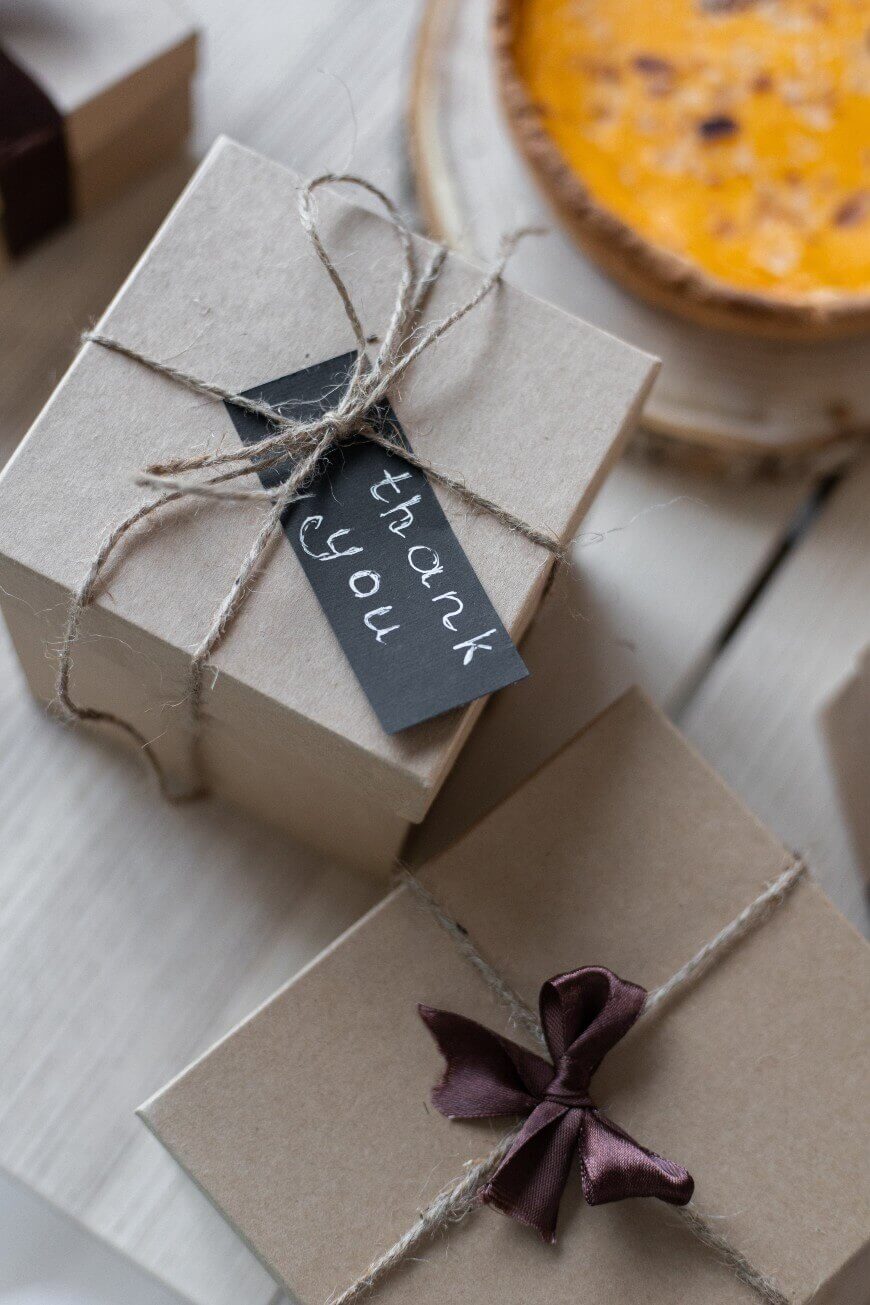 A close-up of two wrapped gift boxes, one tied with twine and a thank you tag, the other with a dark brown ribbon. A pie is partially visible in the background on a light wooden surface.
