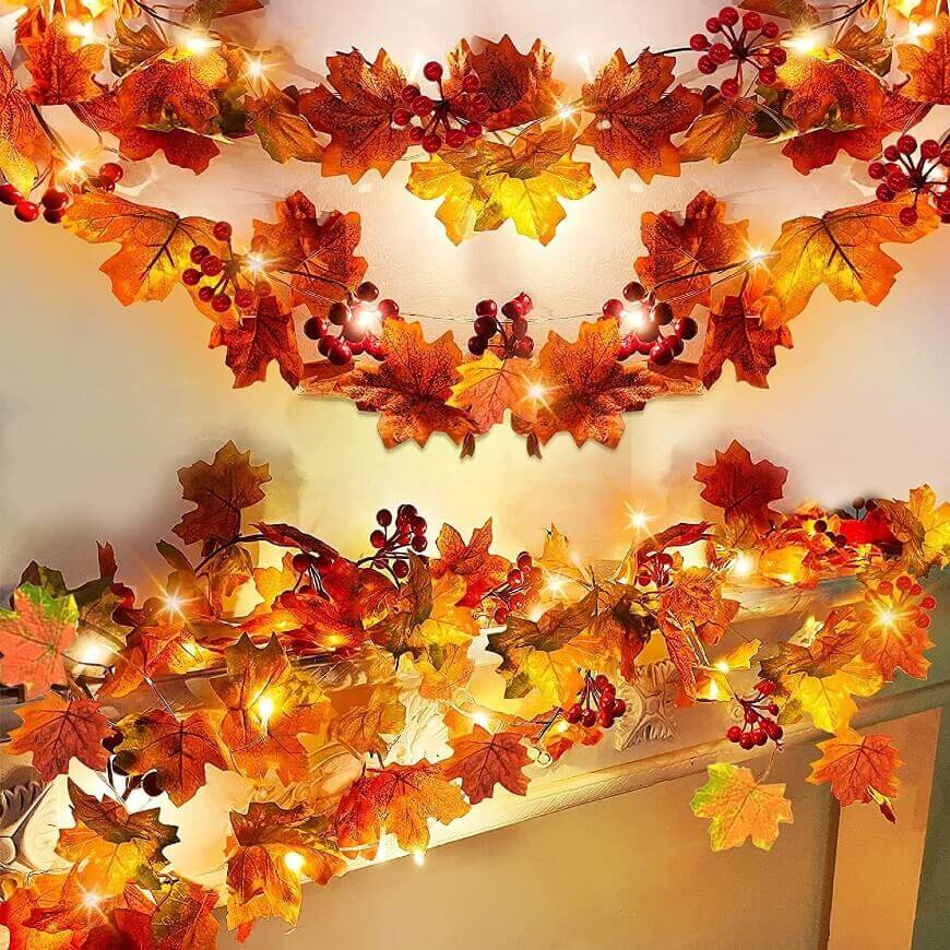 String lights decorated with artificial autumn leaves and red berries, glowing warmly and draped along a mantel and wall, creating a festive fall-themed display.
