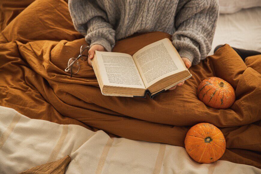 A person wearing a gray sweater sits on a brown blanket, holding an open book. Nearby, a pair of eyeglasses and two small decorative pumpkins rest on the blanket.