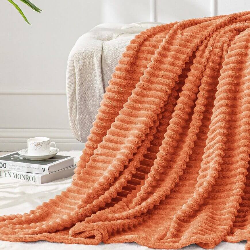 A soft, orange, ribbed blanket is draped over a white sofa next to a stack of books and a teacup on a saucer.