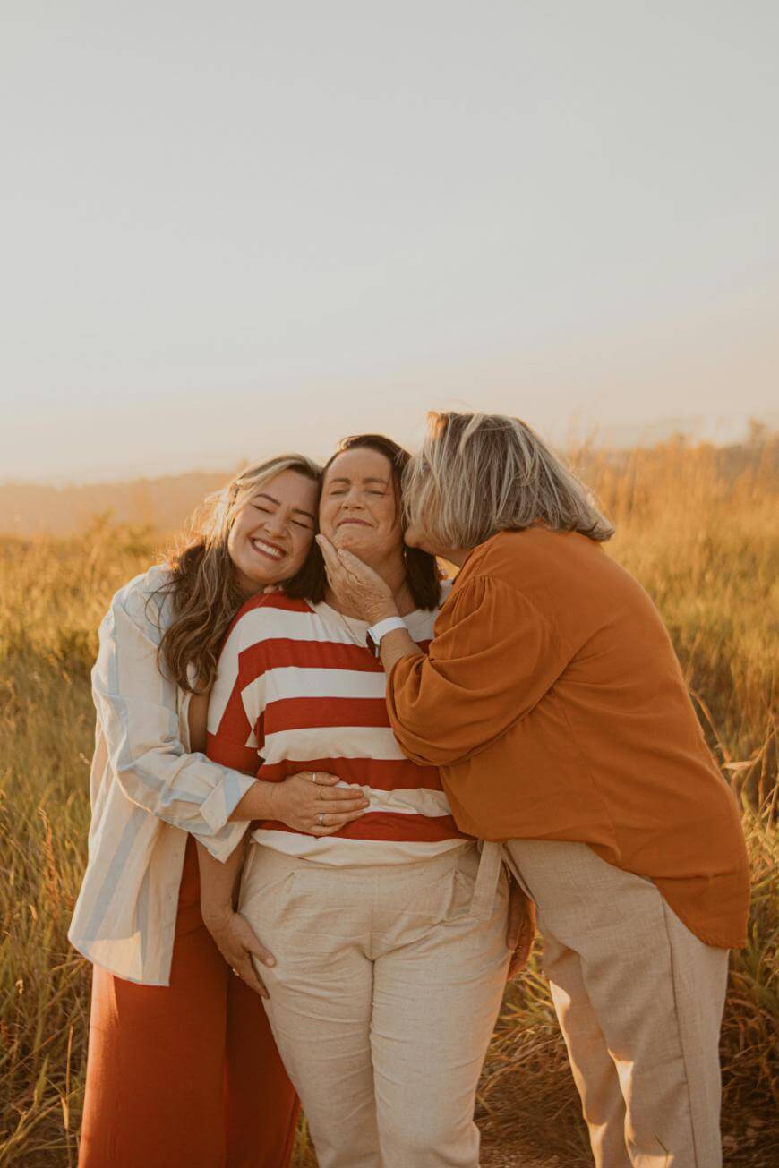 Three women stand close together in a sunlit field, smiling and laughing. One woman in a striped shirt is in the center, while the others embrace her affectionately, radiating warmth and happiness.