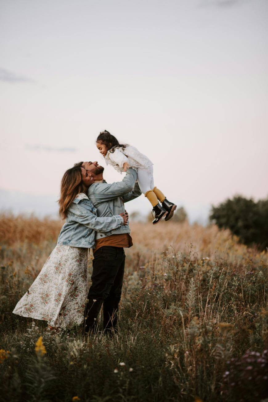 A couple stands in a grassy field at sunset, smiling as they lift a young girl in the air. The woman wears a floral skirt and denim jacket; the man wears dark clothes. The girl is in a white dress and yellow tights.
