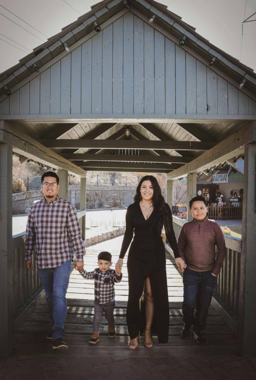 A family of four—two adults and two young boys—hold hands and smile while standing under a wooden covered bridge on a sunny day. The adults are in the center, flanked by the children on each side.
