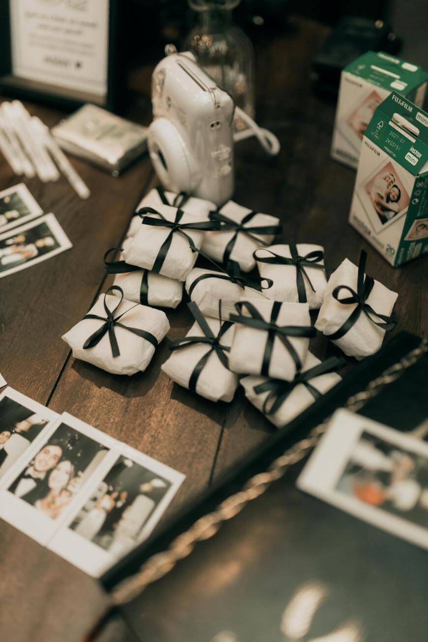 Small gifts wrapped in white paper with black ribbons, instant photos, a white instant camera, and film boxes are arranged on a wooden table, creating a festive and nostalgic atmosphere.
