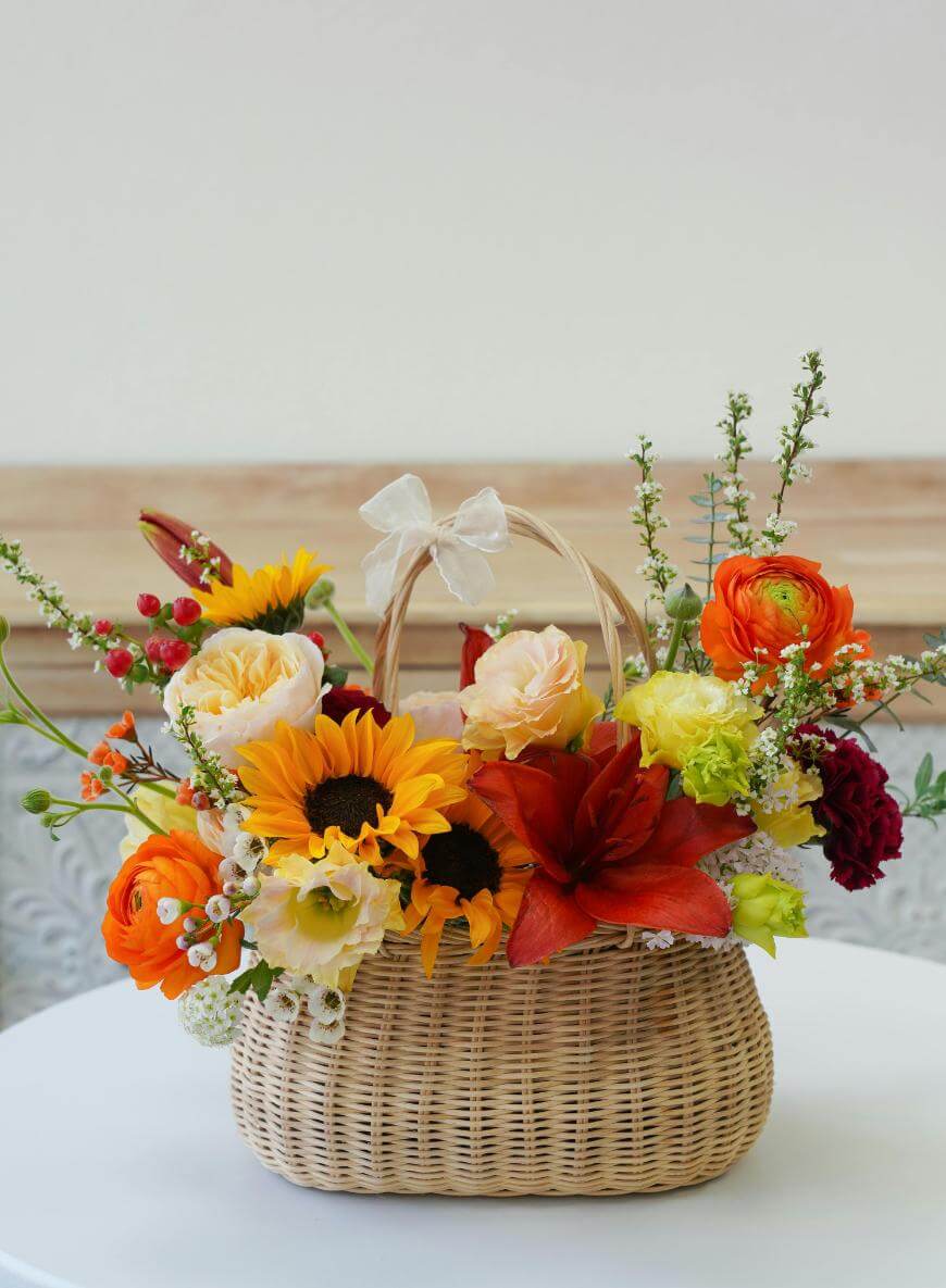 A woven basket filled with a colorful arrangement of sunflowers, roses, lilies, and assorted flowers sits on a white table. The basket has a handle with a small white bow on top.