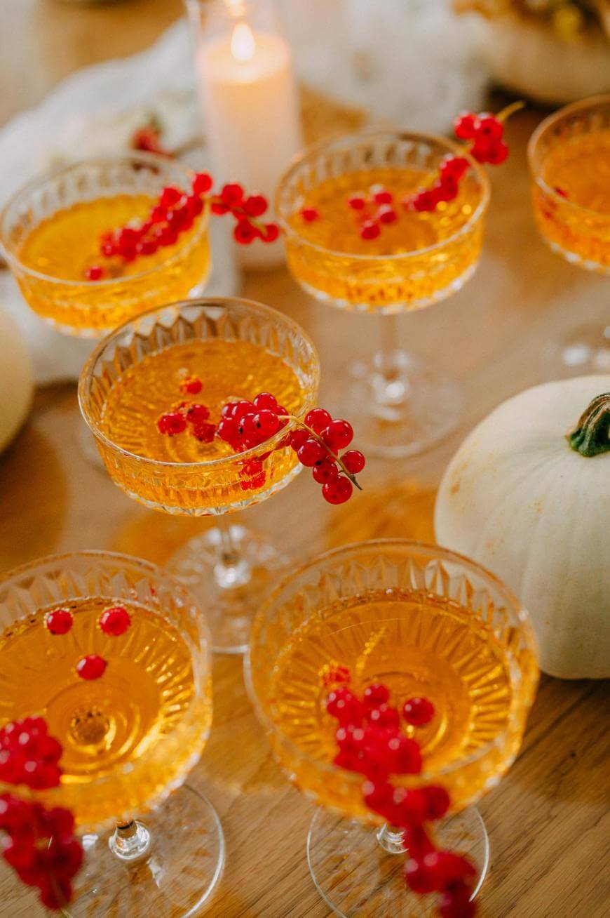 Elegant coupe glasses filled with a golden-orange drink, each garnished with a sprig of red currants. A lit candle and white pumpkins are visible in the background, creating a festive atmosphere.