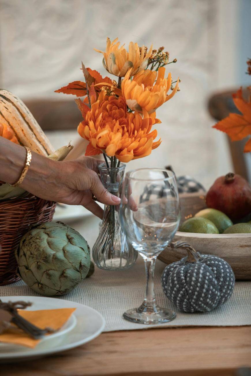 A hand arranges orange flowers in a vase on a table set with a wine glass, an artichoke, fabric pumpkin, fruit bowl, and a plate with cutlery and a yellow napkin. Autumn decor is visible throughout.