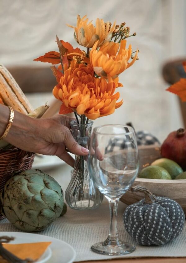 A hand places a vase of orange flowers on a table set with a wine glass, artichoke, fabric pumpkin, squash, and a bowl of fruit, creating an autumn-themed arrangement.
