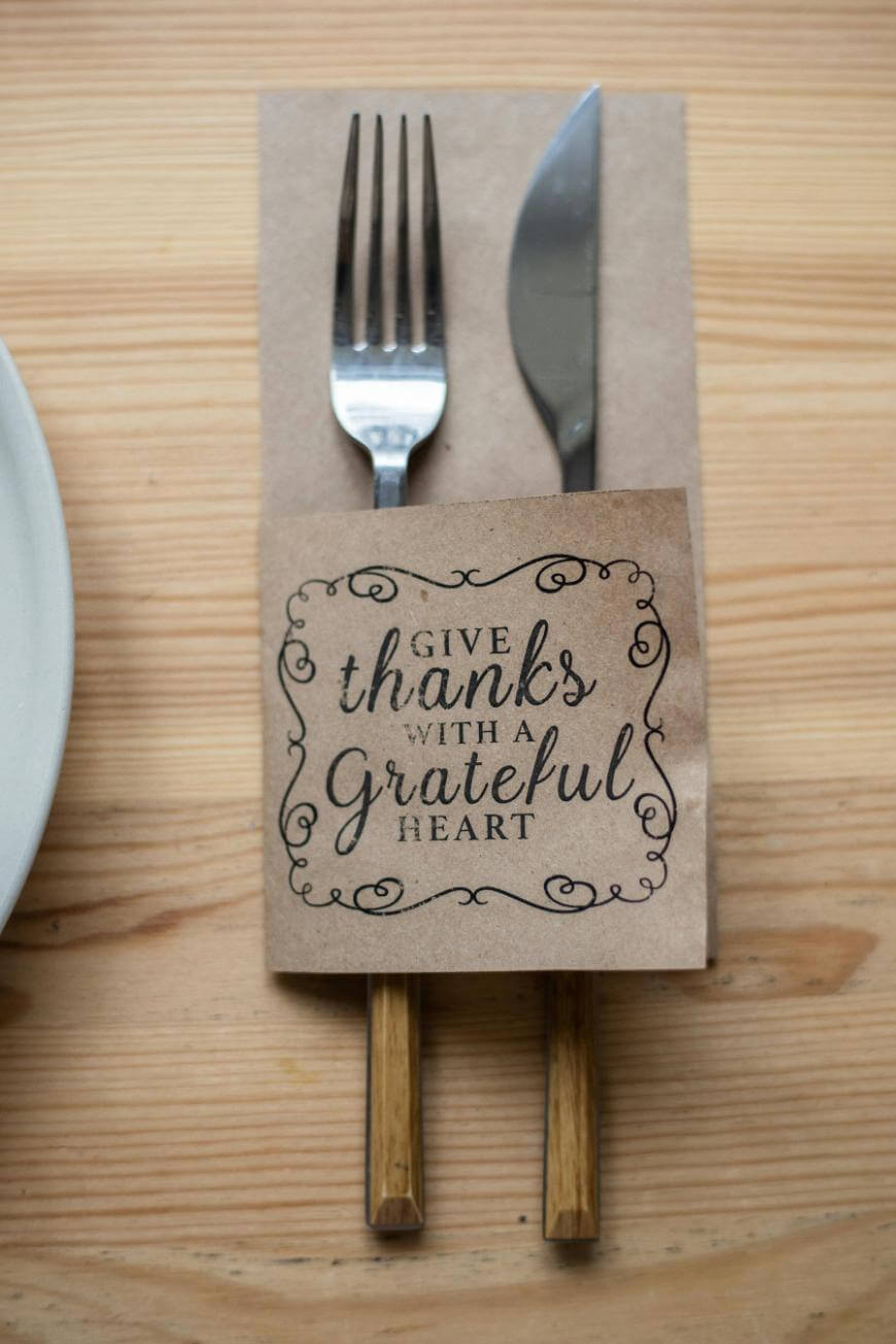 A fork and knife rest on a napkin with a brown paper holder that reads Give thanks with a grateful heart, placed on a wooden table next to a white plate.