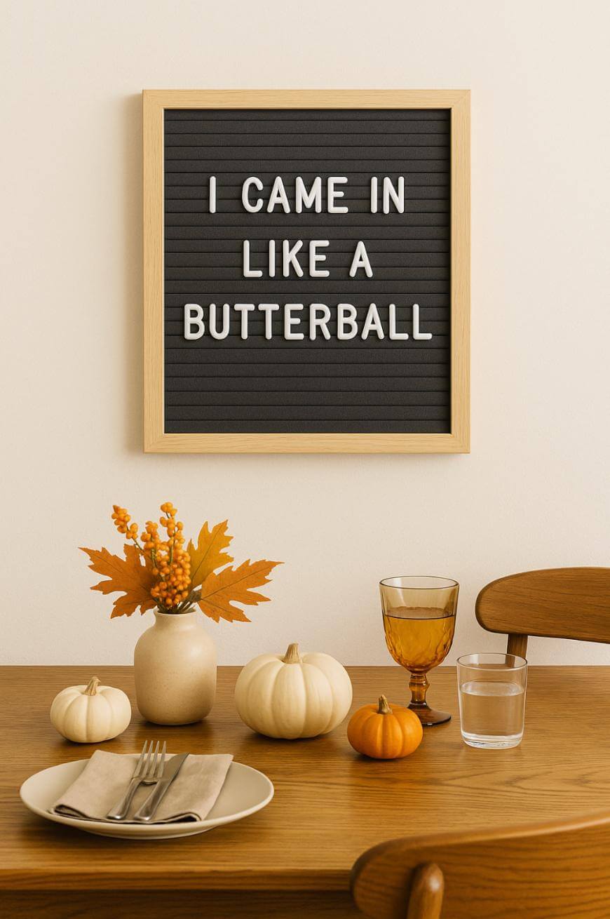 A letter board on a wall reads “I CAME IN LIKE A BUTTERBALL.” Below, a dining table is set with plates, napkins, glasses, small pumpkins, and a vase with autumn leaves.