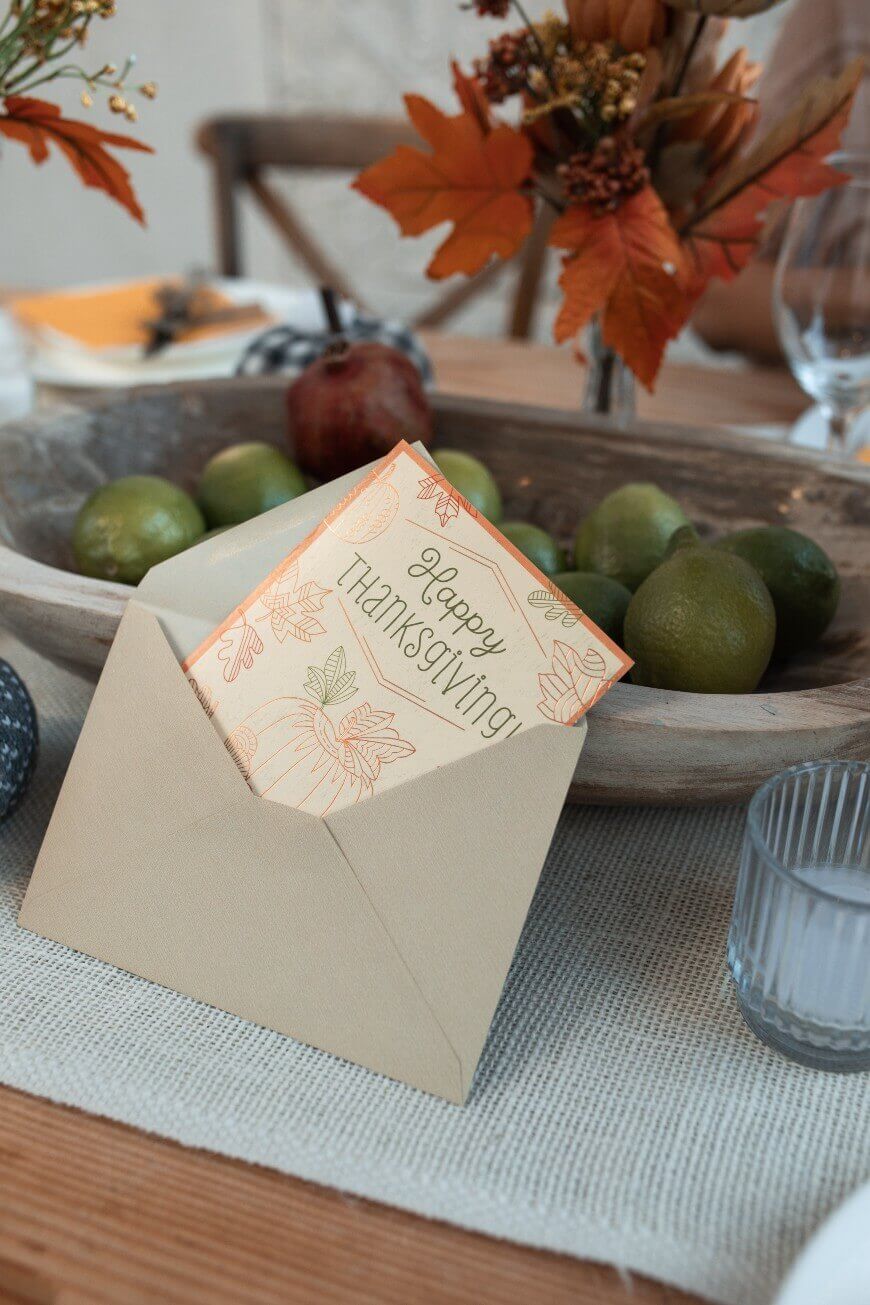 A beige envelope with a “Happy Thanksgiving!” card rests on a table next to a wooden bowl filled with limes. Autumn leaves and tableware can be seen in the background.