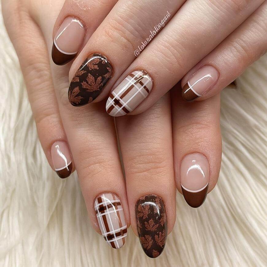 A hand with fall-themed nail art featuring brown French tips, brown and white plaid patterns, and dark brown nails adorned with copper leaf designs, displayed against a soft, cream-colored background.